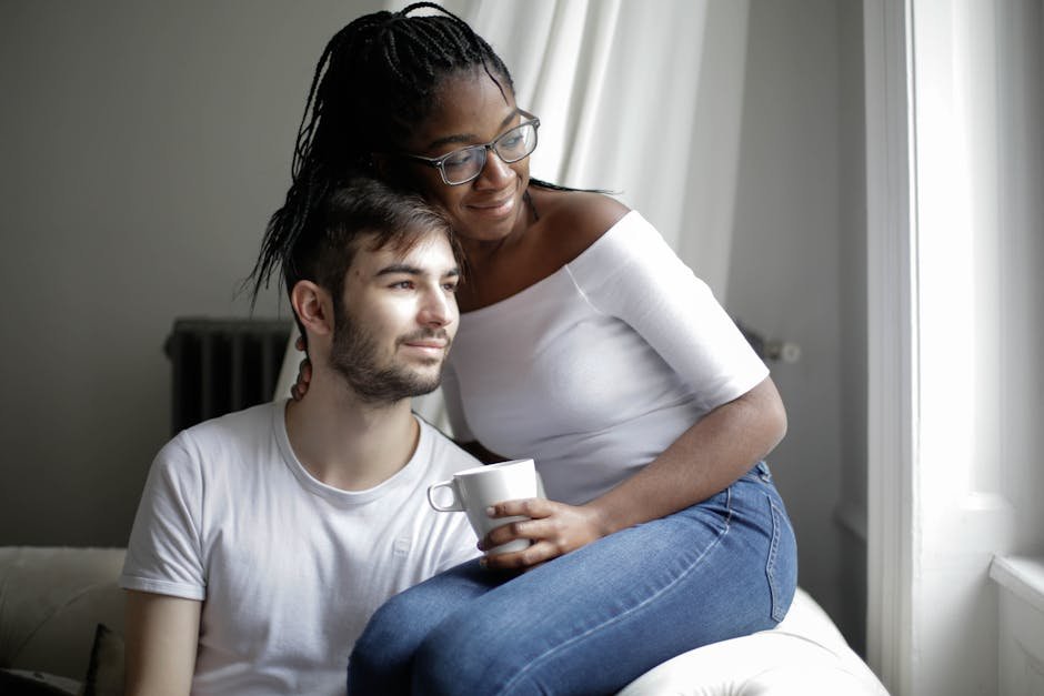 Positive young multiracial couple in casual clothes sitting on comfortable sofa with cup and looking away through window in modern living room, financial challenges